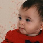 Adorable baby girl with earrings in a red dress, smiling indoors.